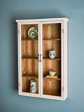 A white wooden wall-mounted cabinet with two glass doors and four interior wooden shelves, styled with ceramic dishes and a pitcher against a muted blue-gray wall.