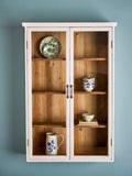 A tall white wooden display cabinet with two glass doors and four interior wooden shelves, styled with ceramic dishes and a pitcher against a muted blue-gray wall.