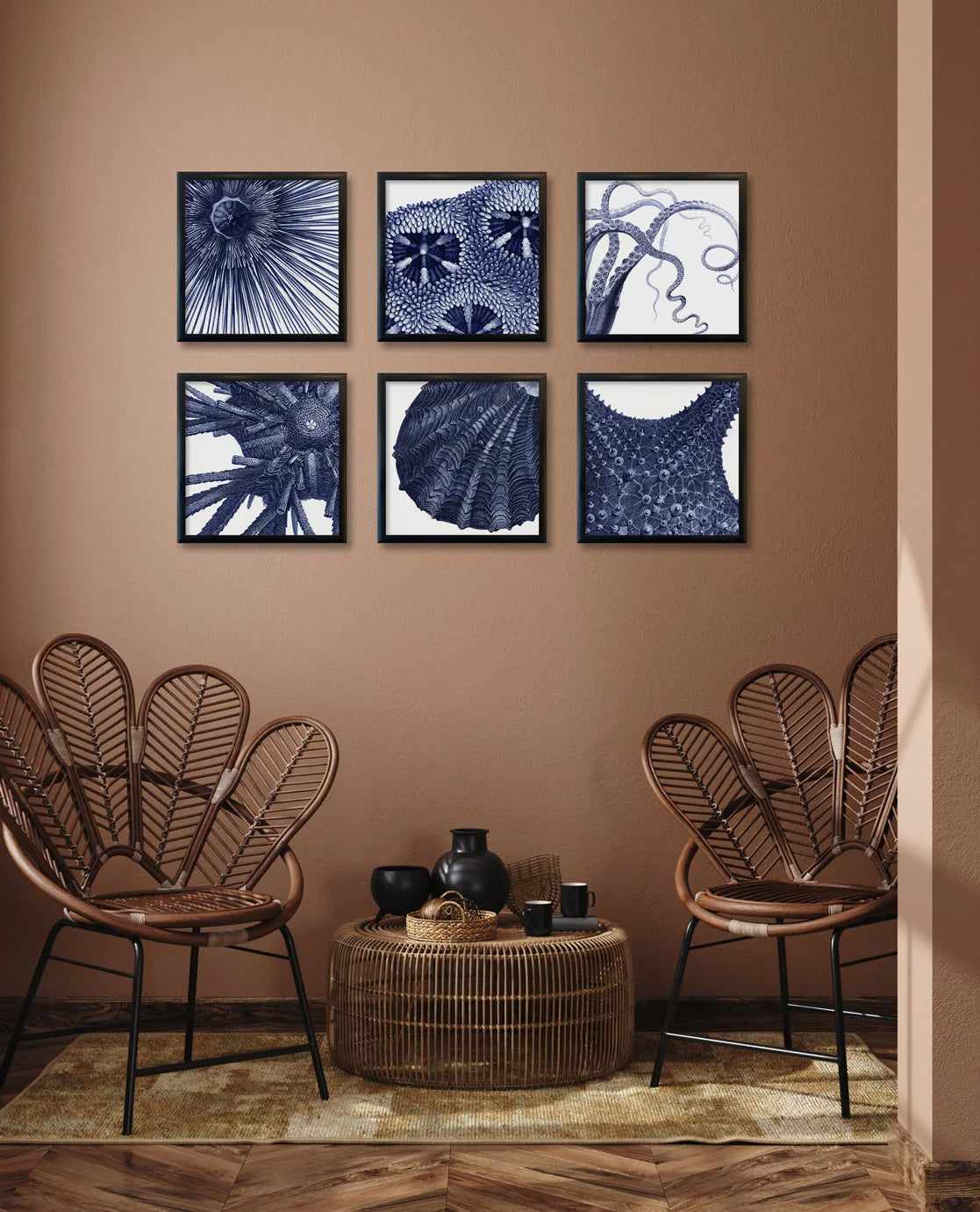A styled interior scene with a warm brown wall, six framed abstract navy-and-white prints arranged in a grid, two woven fan-back chairs, and a round wicker coffee table holding a vase and cups.