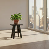 A rustic black wooden stool with a square seat and four angled legs connected by a horizontal stretcher, shown against a plain white background.