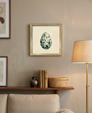 A framed art print featuring a speckled blue-green egg on a cream background, hanging on a textured beige wall above a wooden console shelf with books, a vase, and a basket, next to a floor lamp.