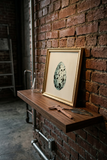 A framed art print of a speckled egg in a gold frame, styled on a dark wooden shelf against a red brick wall.