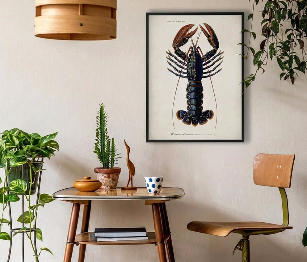 A framed lobster art print hangs on a beige wall in a styled room with a wooden chair, a small table, and several green houseplants.