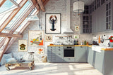 A bright, modern loft kitchen with gray cabinets, white brick walls, exposed wood beams, and framed art on the wall. A small wooden cart with cushions sits near the window, and the kitchen is well-lit with natural light from the skylight.