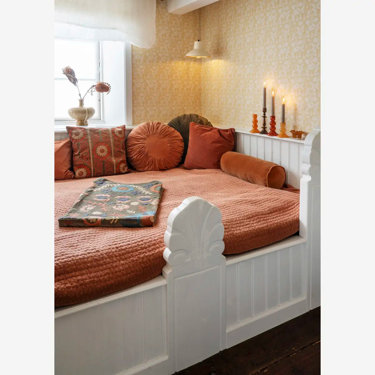 A white wooden daybed with a textured rust-orange quilt and matching pillows, styled in a cozy room with patterned wallpaper, candles, and a window.