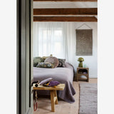 A cozy bedroom interior with a bed dressed in gray bedding, patterned pillows, a small wooden side table with a green lamp, a hanging wall textile, exposed wooden ceiling beams, and a light gray rug.