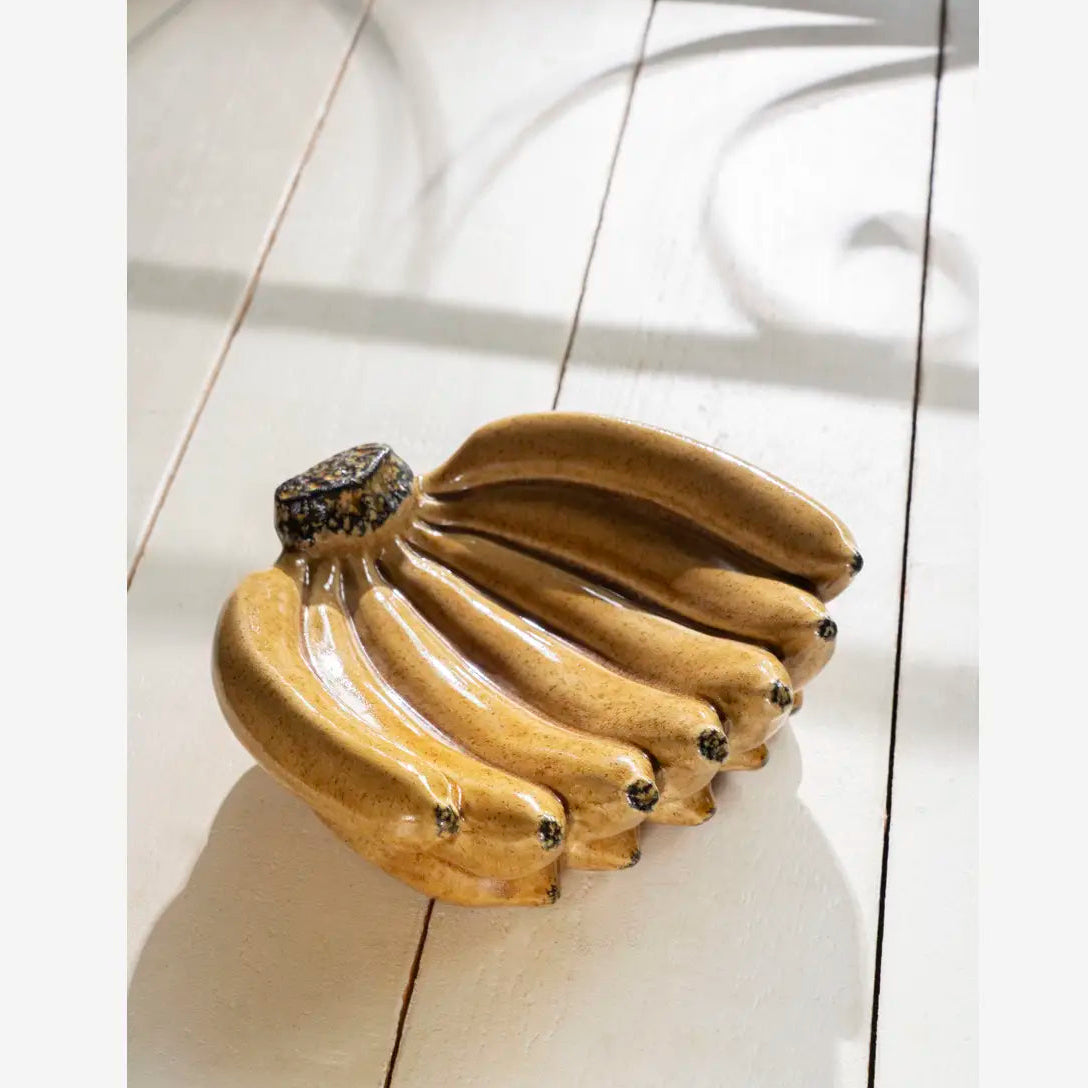 A ceramic sculptural figurine shaped like a cluster of bananas, displayed on a white wooden plank surface with soft shadows.