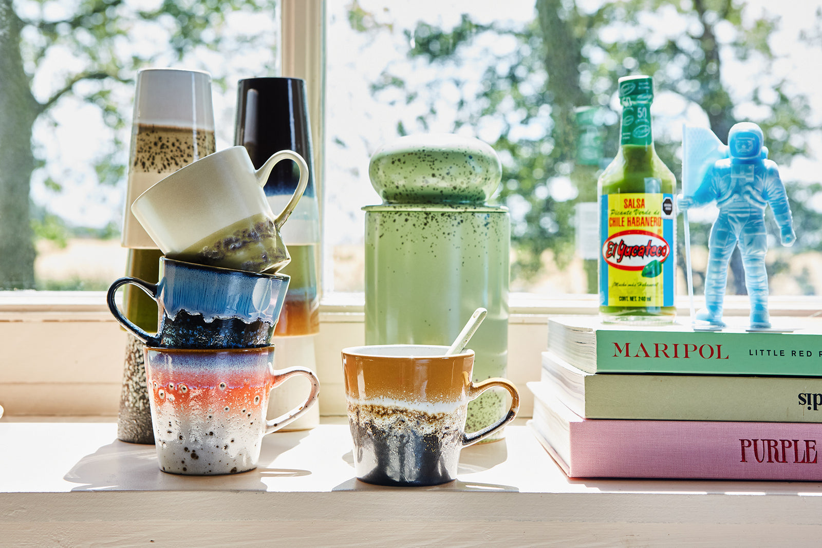 Colorful mugs and books on a windowsill with a view of trees.