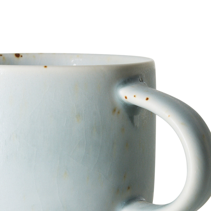 Close-up of a ceramic mug with a pale blue-gray glaze and small brown speckled distressing, showing the side handle against a white background.