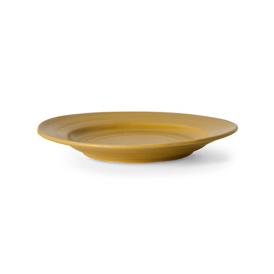 A wide, shallow round plate in a matte mustard yellow color, shown from a side angle on a clean white background.