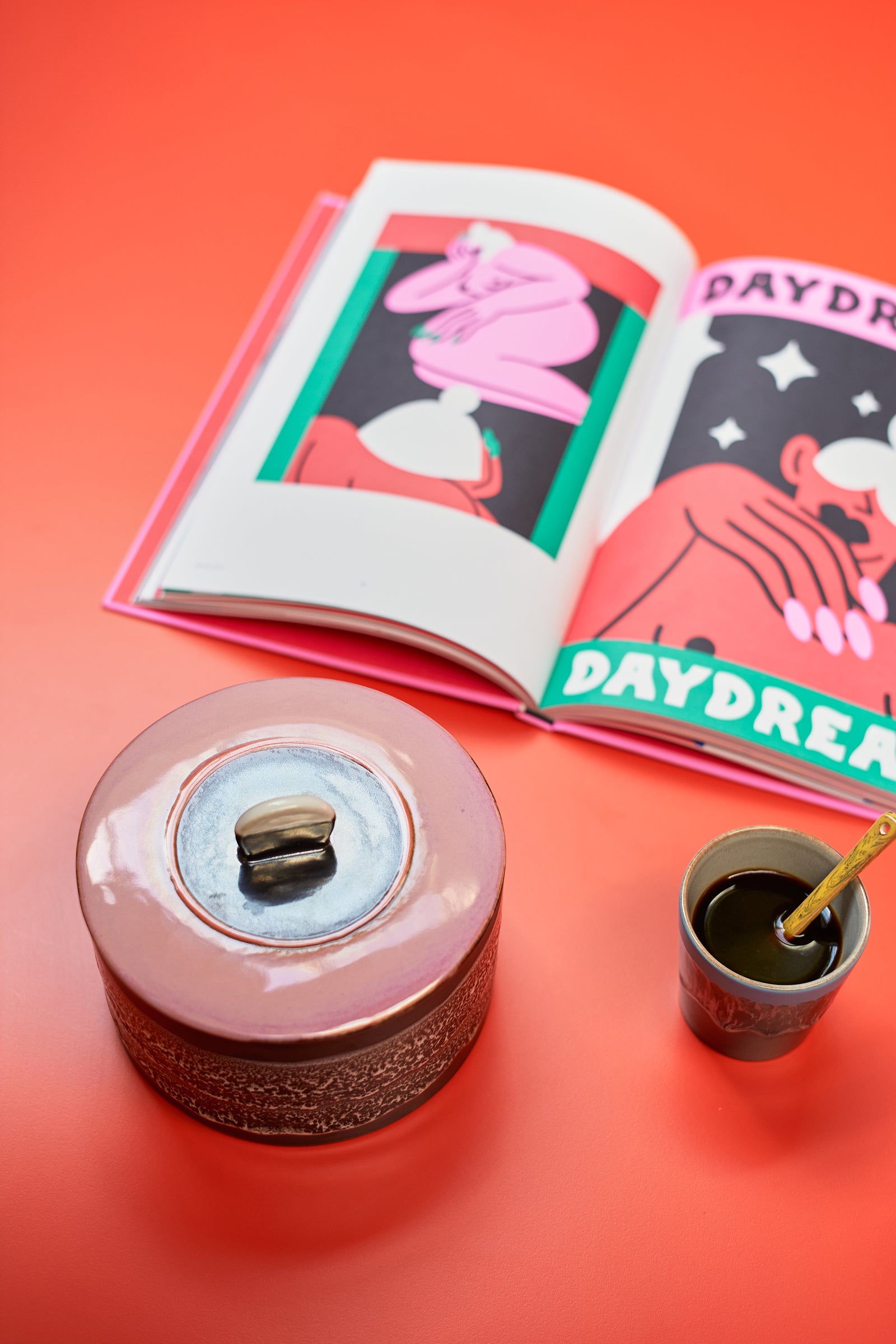 Ceramic container with a lid, small cup with straw, and open book titled 'Daydream' on a red background